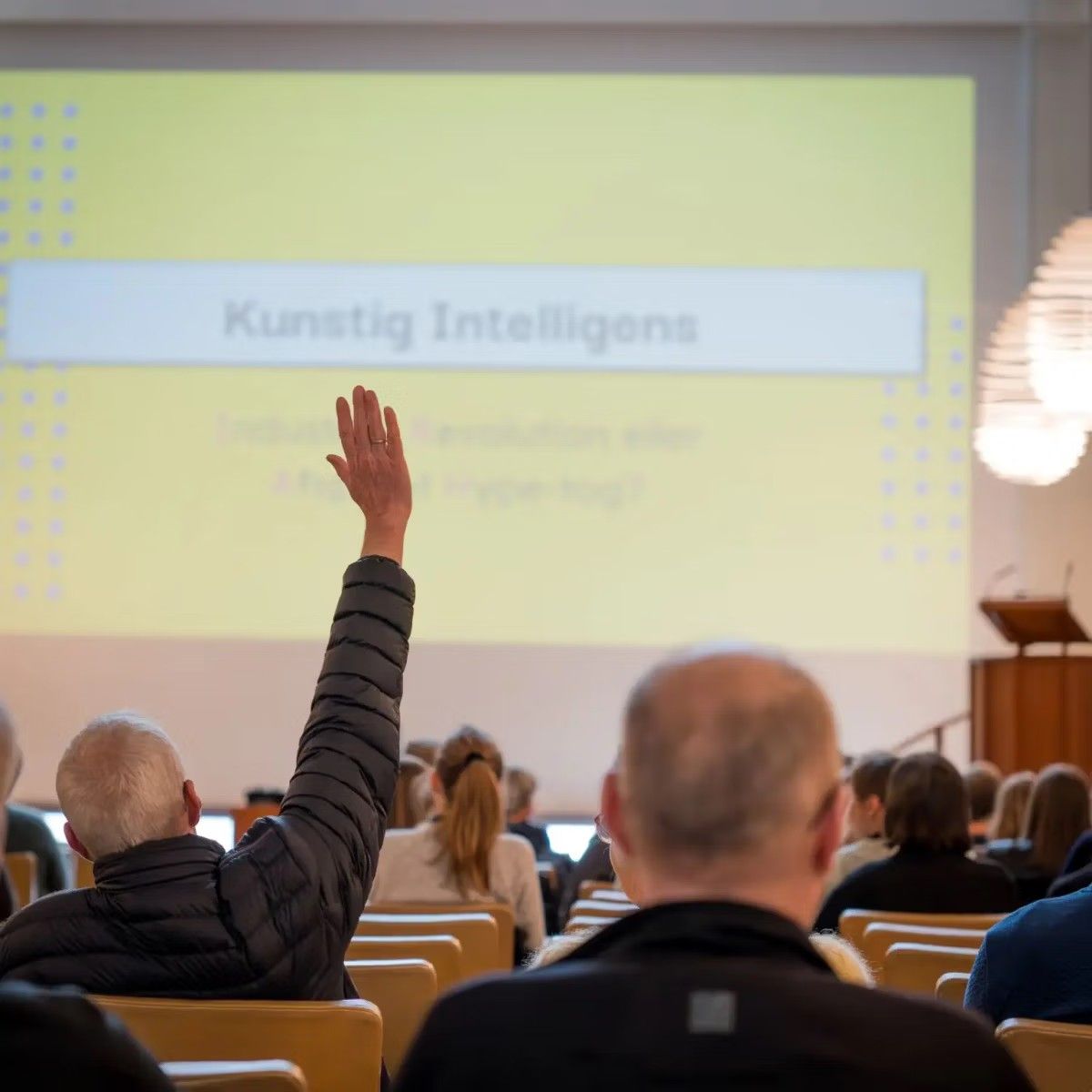 The audience at Aarhus BSS seen from behind, one person raising a hand towards a yellow slide that reads Kunstig Intelligens