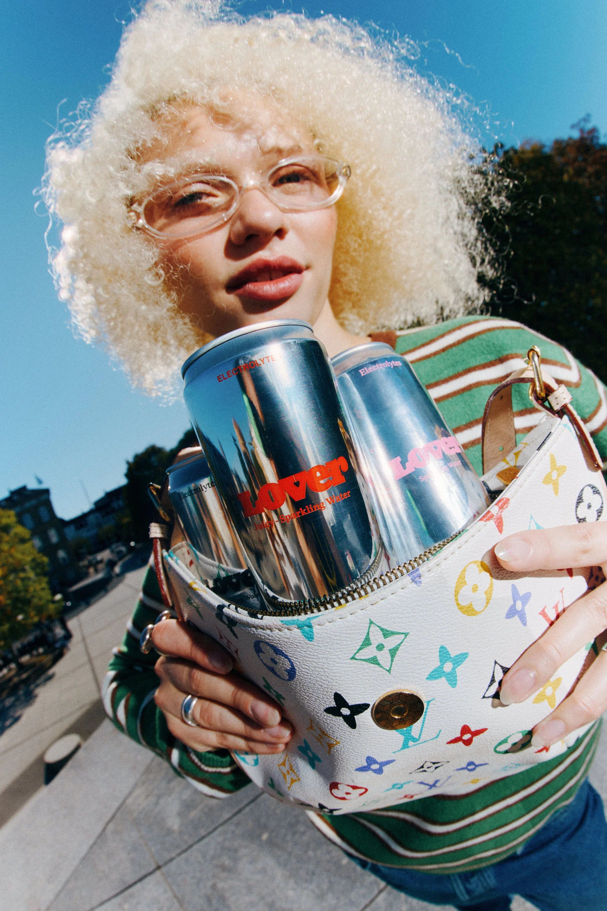 A woman with blonde curls and clear glasses holds out a colourful monogram handbag stuffed with chrome Lover cans
