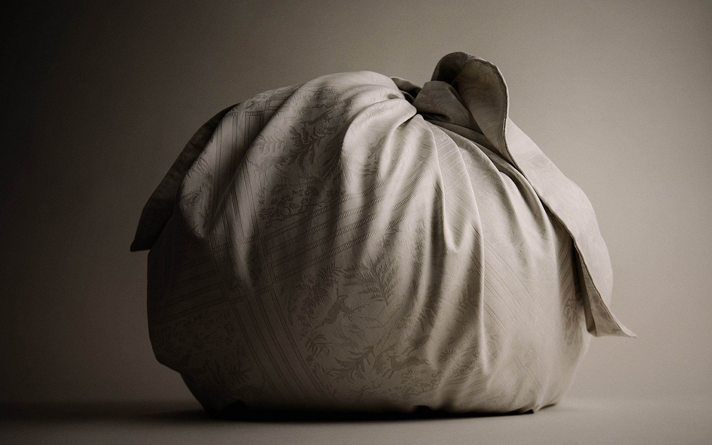 A bundle of Avillafranciska jacquard bedding tied into a soft round form, lit low against a dark ground
