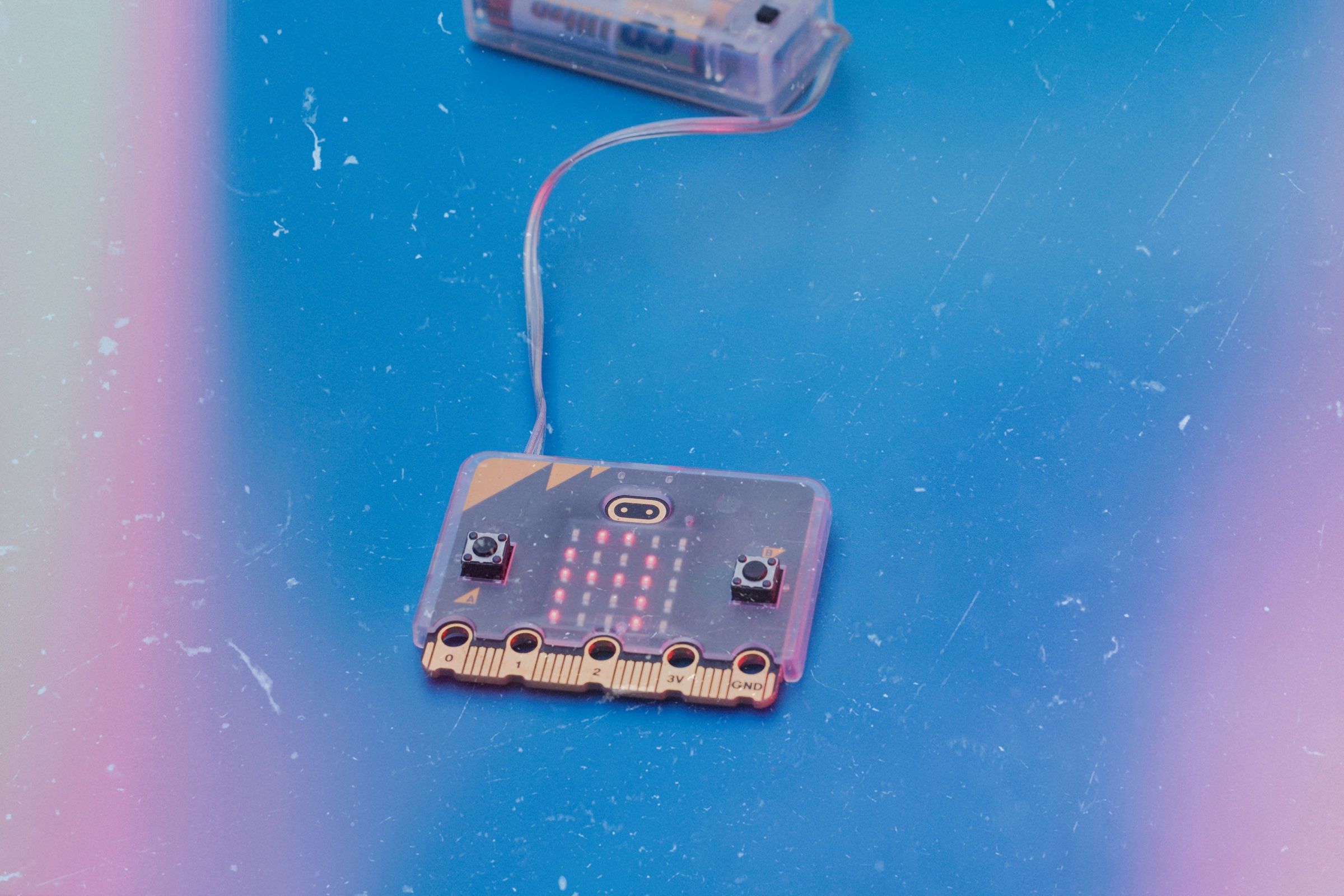 A BBC micro:bit with its red LED grid lit, wired to a battery pack on a blue table