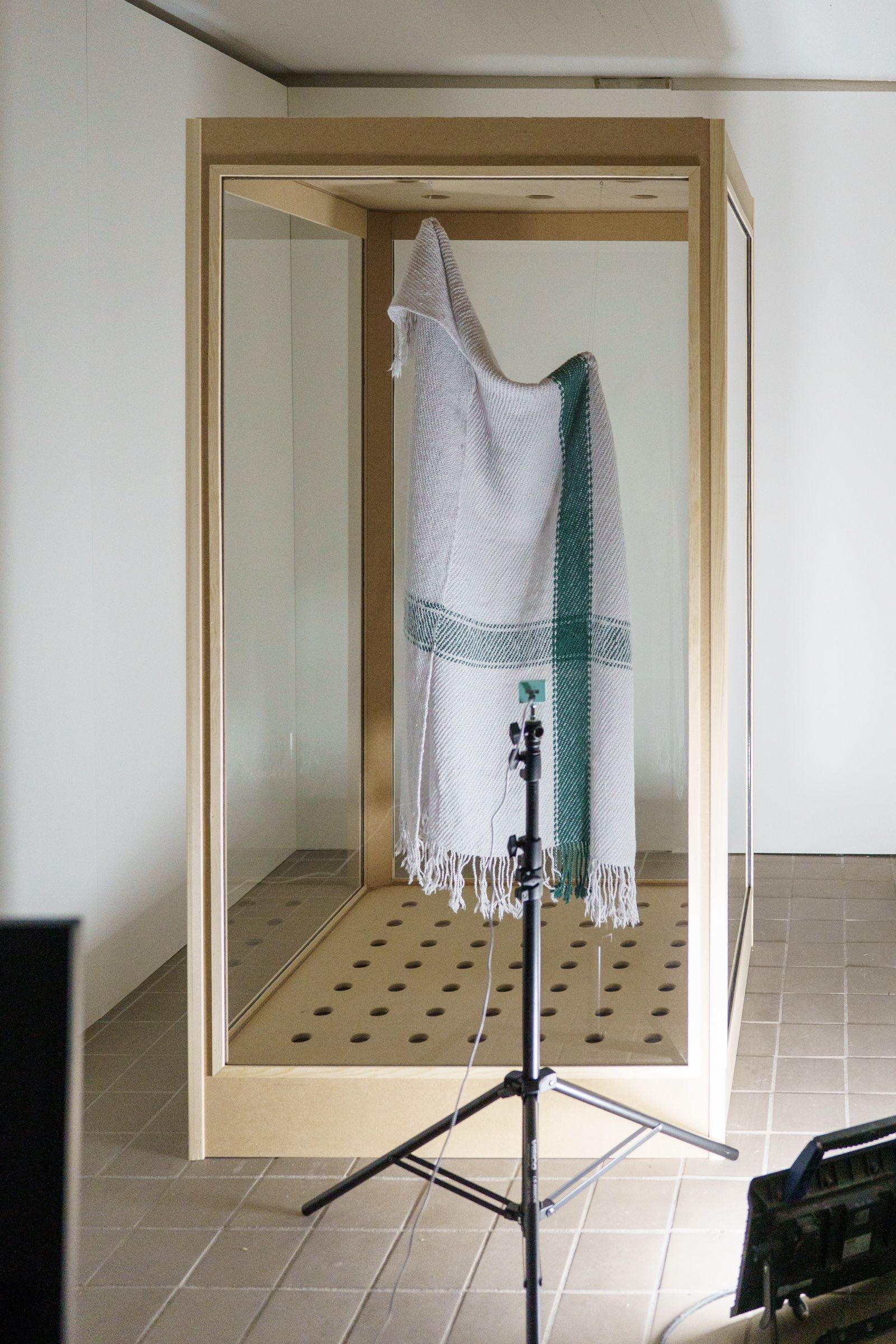 The installation, a cream plaid with green stripes hanging in a tall wood and glass cabinet with a perforated floor, a camera on a stand in front